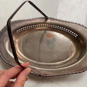 May include: Silver-toned oval serving basket with a handle. The basket has a decorative pierced design around the rim and a handle that arches over the top. The basket is likely made of metal and has a reflective surface.