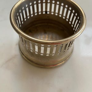 Op de afbeelding: Een vintage messing onderzetter met een decoratief opengewerkt ontwerp. De onderzetter heeft een cirkelvormige vorm met een licht uitlopende basis en een reeks verticale sleuven aan de zijkanten. Het messing heeft een warme, verouderde patina.