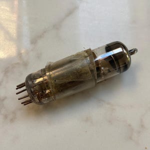 Könnte beinhalten: Eine Vintage-Vakuumröhre mit einem klaren Glaskörper und einem Metallsockel. Die Röhre ist mit braunem Papierklebeband umwickelt und hat neun Stifte am Sockel.