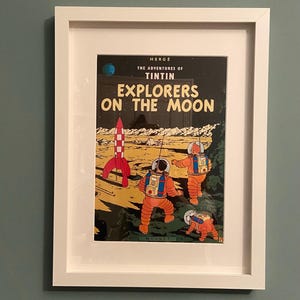 May include: Framed print of a comic book cover titled "Explorers on the Moon" by Hergé. The artwork features astronauts in orange suits on a lunar landscape with a red rocket. The print is in a white frame.