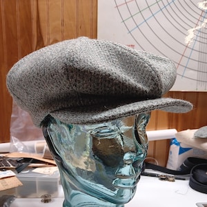 May include: A grey tweed newsboy cap on a glass mannequin head. The cap features a rounded crown and a short, stiff peak. The cap is a neutral grey colour and appears to be made of a textured fabric. The mannequin head is a light blue colour.