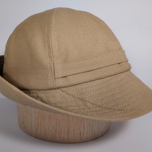 May include: A beige hat with a short brim and a structured crown. The hat has a band detail above the brim and is made of a textured fabric. The hat is displayed on a wooden form, highlighting its shape and design.