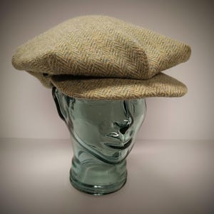 May include: A green and brown tweed flat cap with a herringbone pattern. The cap is on a glass mannequin head.