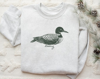 Puede incluir: Una sudadera gris claro con una ilustración detallada de un somorgujo en verde oscuro. La palabra "loony" está escrita en cursiva debajo del pájaro. La sudadera está hecha de un material suave.