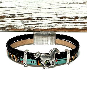 May include: Black braided leather bracelet with a silver horse charm and a turquoise band featuring an arrow design. The bracelet has a magnetic clasp and decorative metal accents.