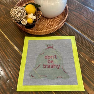 May include: A needlepoint canvas with a green trash bag design and the words "don't be trashy" in pink. The canvas is framed with a bright yellow border. A decorative tray with a white vase and decorative items sits in the background.