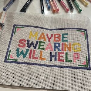 May include: A needlepoint canvas with the phrase "MAYBE SWEARING WILL HELP" in colorful block letters. The canvas is surrounded by a blue border with green and pink accents. Various colored markers are scattered above the canvas.