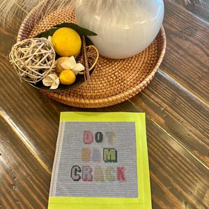 May include: A square needlepoint canvas with the words "DO IT M CRUEL" in colorful letters, framed by a bright yellow border. The canvas is on a wooden surface, with a woven tray, white vase, and decorative items in the background.