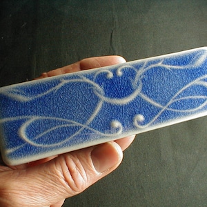 May include: A rectangular ceramic tile with a blue glaze and white swirling designs. The tile has a textured surface.