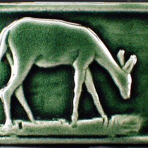 May include: A rectangular, green ceramic tile featuring a relief of a deer grazing. The deer is depicted in a light green color, with its head lowered to the ground. The tile has a textured surface and a dark green border.