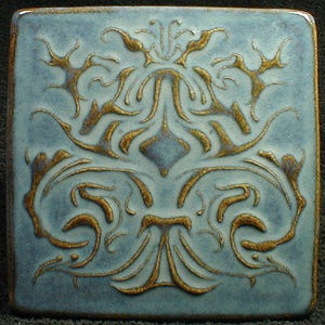 May include: Square ceramic tile with a light blue background and a raised gold floral design. The design is symmetrical and features stylized leaves and curves. The tile has a glossy finish and a dark brown border.
