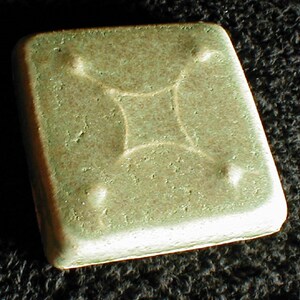 May include: A square, light green ceramic tile with a raised, four-pointed star design in the center. The tile has rounded edges and small, raised dots at each corner of the star. The surface has a textured appearance.