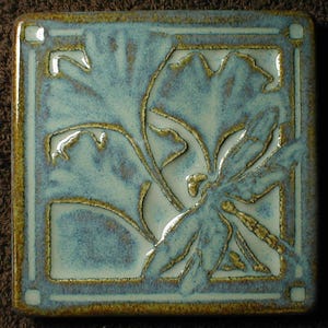 May include: A ceramic tile with a dragonfly and floral design. The dragonfly is outlined in gold and the background is a light blue-green color.