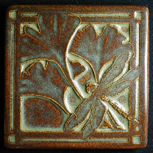 May include: Square ceramic tile with a dragonfly and leaf design. The tile features a raised relief pattern in shades of brown and green, with a square border. The dragonfly is centered, with detailed wings and body.