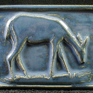 May include: A rectangular ceramic tile with a raised relief of a deer in a field. The tile has a blue glaze with gold accents around the edges and on the deer's legs and antlers. The deer is depicted grazing.