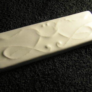 May include: A white ceramic tile with a raised design of stylized leaves and swirls.