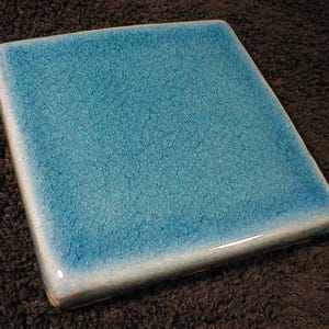 May include: Square ceramic tile with a crackle glaze in shades of turquoise and blue. The tile has a glossy finish and a white border. The tile is a decorative element.