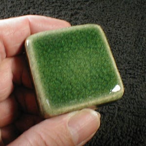 May include: A square, ceramic tile with a textured, dark green surface and a light brown border. The tile is held in a hand, showcasing its glossy finish and unique color.