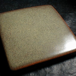 May include: A square, ceramic box with a speckled, light green surface and rounded edges. The edges are a darker brown color. The box has a glossy finish and appears to be handmade.