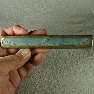 May include: A rectangular ceramic piece with a light blue center and gold trim. The design includes a gold line and a small gold emblem. The object is held in a hand.