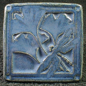 May include: Square ceramic tile with a dragonfly and leaf design. The tile is a muted blue with raised, textured details in a lighter, silvery color. The dragonfly is centered, with leaves in the background, all within a square border.