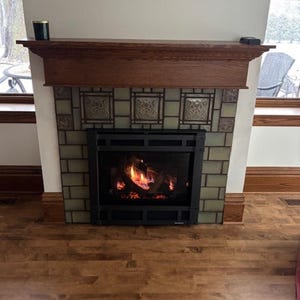 May include: A fireplace with a dark wooden mantel and a black metal firebox. The firebox contains a glowing fire. The fireplace is surrounded by green and patterned tiles. The floor is made of wooden planks.