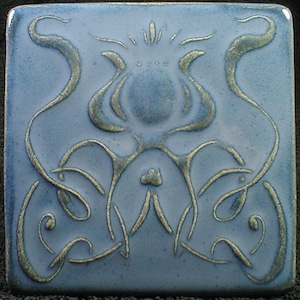 May include: Square ceramic tile with a light blue background and a raised, stylized floral design in a metallic gold color. The design features flowing lines and a central flower-like motif, creating an Art Nouveau aesthetic.