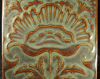 May include: Square ceramic tile with a floral design. The tile features a central flower-like motif with radiating lines and stylized leaves, all in a warm brown glaze against a muted green background. The edges are also brown.
