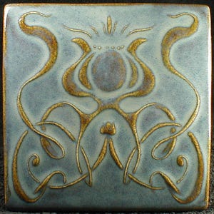 May include: Square ceramic tile with a light blue background and a gold floral design. The design features stylized flower and vine motifs. The tile has a glossy finish and a dark border.