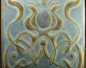 May include: Square ceramic tile with a light blue background and a gold floral design. The design features stylized flower and vine motifs. The tile has a glossy finish and a dark border.