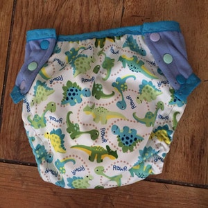 May include: A white cloth diaper with a blue trim and a dinosaur print. The dinosaurs are green, blue, and yellow and say "Roar" in a circle around them.