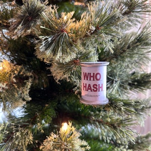 May include: A cylindrical Christmas tree ornament with the words "WHO HASH" in red letters. The ornament is silver and white, hanging on a green Christmas tree with white lights.