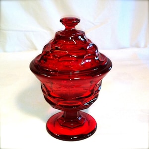 May include: A red glass candy dish with a lid. The dish has a pedestal base and a decorative pattern.