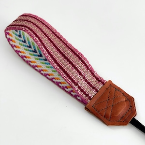 May include: A pink and white camera strap with a colorful geometric pattern and gold glitter accents. The strap has a brown leather end piece.