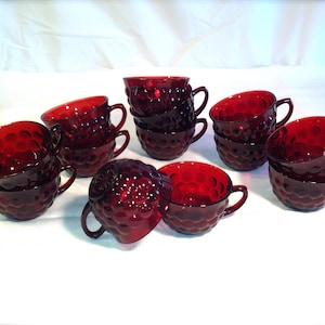 May include: A collection of ruby red glass teacups with a textured, hobnail design. Each cup has a handle and is stacked in sets. The cups are arranged on a white surface, showcasing their vibrant color and classic style.