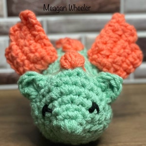 May include: A crocheted green and orange dragon toy with black eyes. The dragon has large orange wings and a small orange horn on its head.
