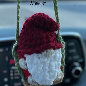 May include: A crocheted gnome ornament hanging from a green string. The gnome has a red hat, white beard, and brown accents. The text "Meagan Wheeler" is visible above the gnome.