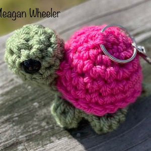 May include: A crocheted turtle keychain with a pink shell and green head and legs. The turtle has black eyes and a silver key ring.