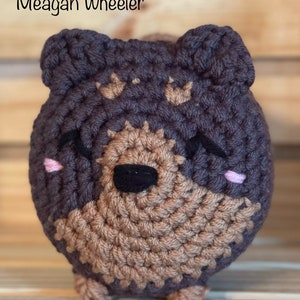 May include: A crocheted plush toy shaped like a brown and gray dog with pink cheeks and black eyes. The toy is standing on a wooden surface.