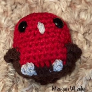 May include: A crocheted red bird with black eyes and gray feet. The bird is sitting on a brown surface.