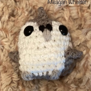 May include: A crocheted plush toy shaped like a penguin with a gray and white body and black eyes. The toy is sitting on a brown fuzzy surface.