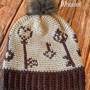 May include: A knitted winter hat with a brown and beige key pattern and a faux fur pom-pom. The hat has a ribbed brown brim and the text "Meagan Wheeler".