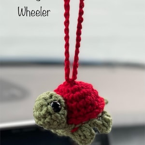 May include: A red and green crocheted turtle hanging from a red string. The turtle has black eyes and is a car charm. The text "Meagan Wheeler" is visible in the background.