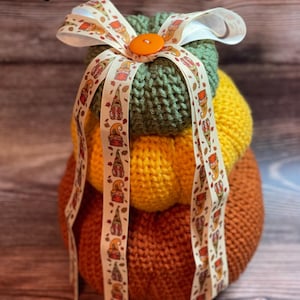 May include: A handmade, stacked pumpkin decoration in shades of orange, yellow, and green. The pumpkin is made of knitted yarn and adorned with a decorative ribbon featuring gnome illustrations. An orange button secures the ribbon. The text "Meagan Wheeler" is visible.
