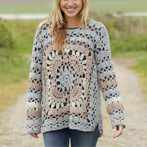 Può includere: Un maglione lavorato a uncinetto con un grande e intricato disegno circolare in tonalità di grigio, beige e bianco. Il maglione ha maniche lunghe e una vestibilità rilassata. Il testo "DROPS DESIGN" e "www.garnstudio.com" è visibile nella parte inferiore dell'immagine.