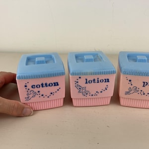 May include: Three square plastic containers with light blue lids and pink bases. Each container features blue text reading cotton, lotion, or pins, accompanied by a graphic of a small cherub surrounded by stars on the front of each unit.