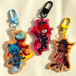 May include: Three colorful acrylic keychains featuring anime-style characters with horns and wings. Each keychain has a different character design, with a blue, red, and brown color scheme. The keychains have a lobster clasp in blue, black, and yellow.