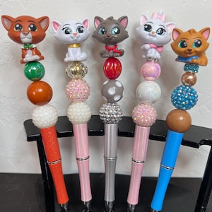 May include: Five decorative pens featuring cat character toppers. The pens have various colors, including orange, pink, silver, and blue. Each pen has a different design of beads and a cat figurine on top. The pens are displayed in a black pen holder.