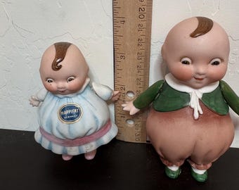 May include: Two vintage ceramic Kewpie dolls. One doll wears a blue dress with the text "HAPPIFAT GERMANY". The other doll wears a green jacket and brown pants. A wooden ruler is placed behind the dolls.