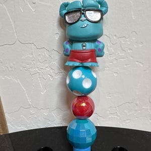 May include: A blue pen with a cartoon character wearing sunglasses, red shorts, and purple polka dots. The pen has a series of decorative beads in blue, red, and blue. The pen is resting in a black pen holder.